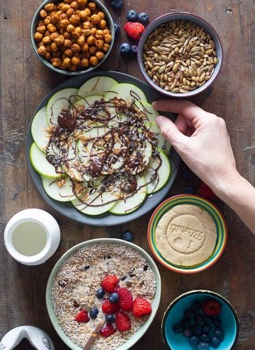 Cereal in a bowl, cookie on a plate, crispy chickpeas in a bowl, seeds in a bowl, all on a wooden table.