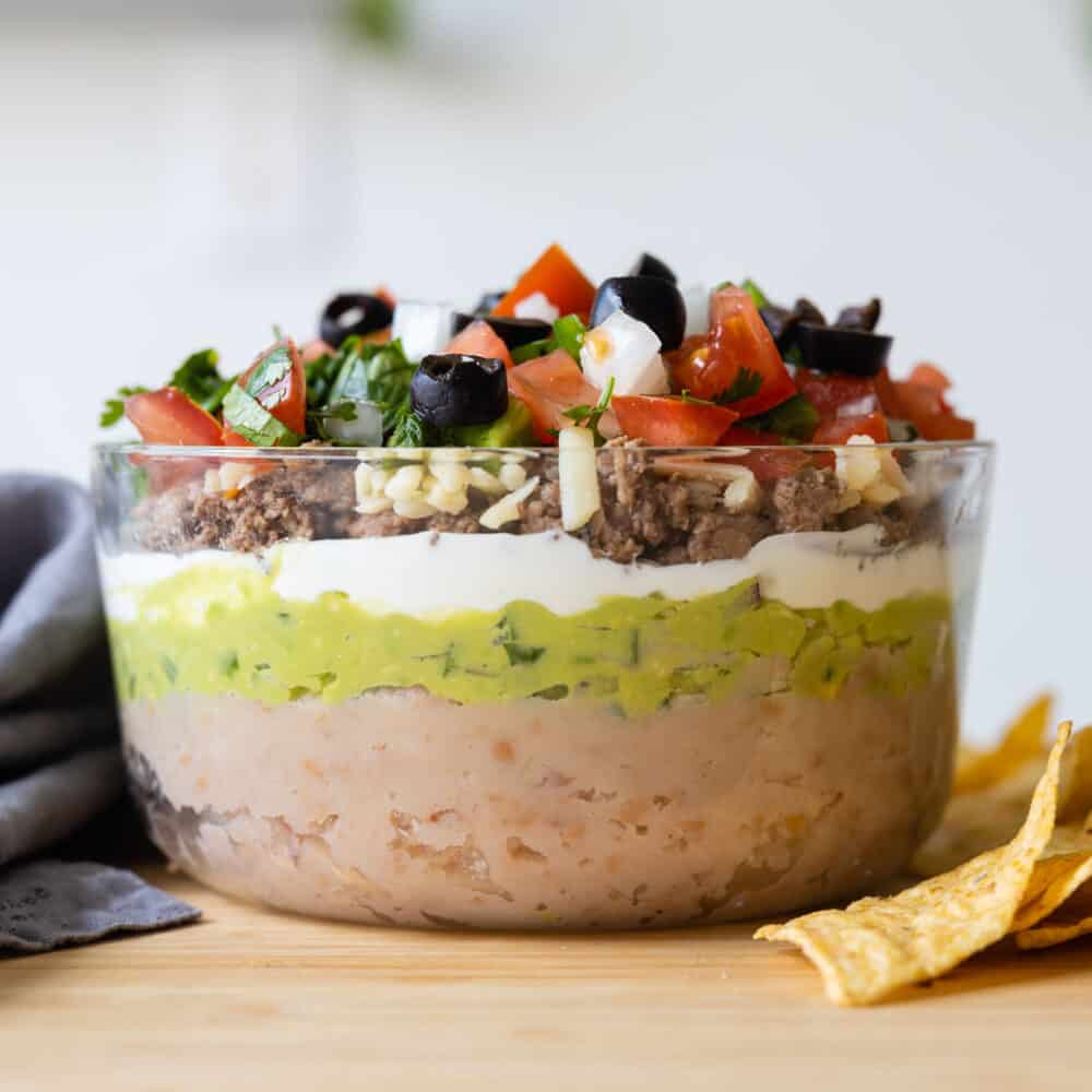 Layers of sophisticated 7-Layer Dip shown at 90 degree angle. Brown beans first, then green guacamole, then white sour cream, then yellow cheese, then brown ground beef, then red white and green pico de gallo, then black olives.