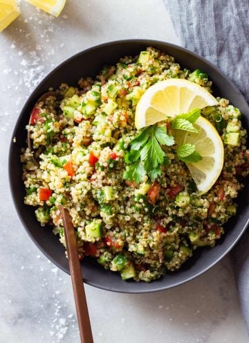 Quinoa Tabbouleh in a gray bowl on a marble surface topped with lemon slices and parsley leaves.