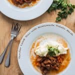 Top view of Tender Lamb Stew with Homemade Potato Mash on white plates on a wooden table with two forks, with text overlay.