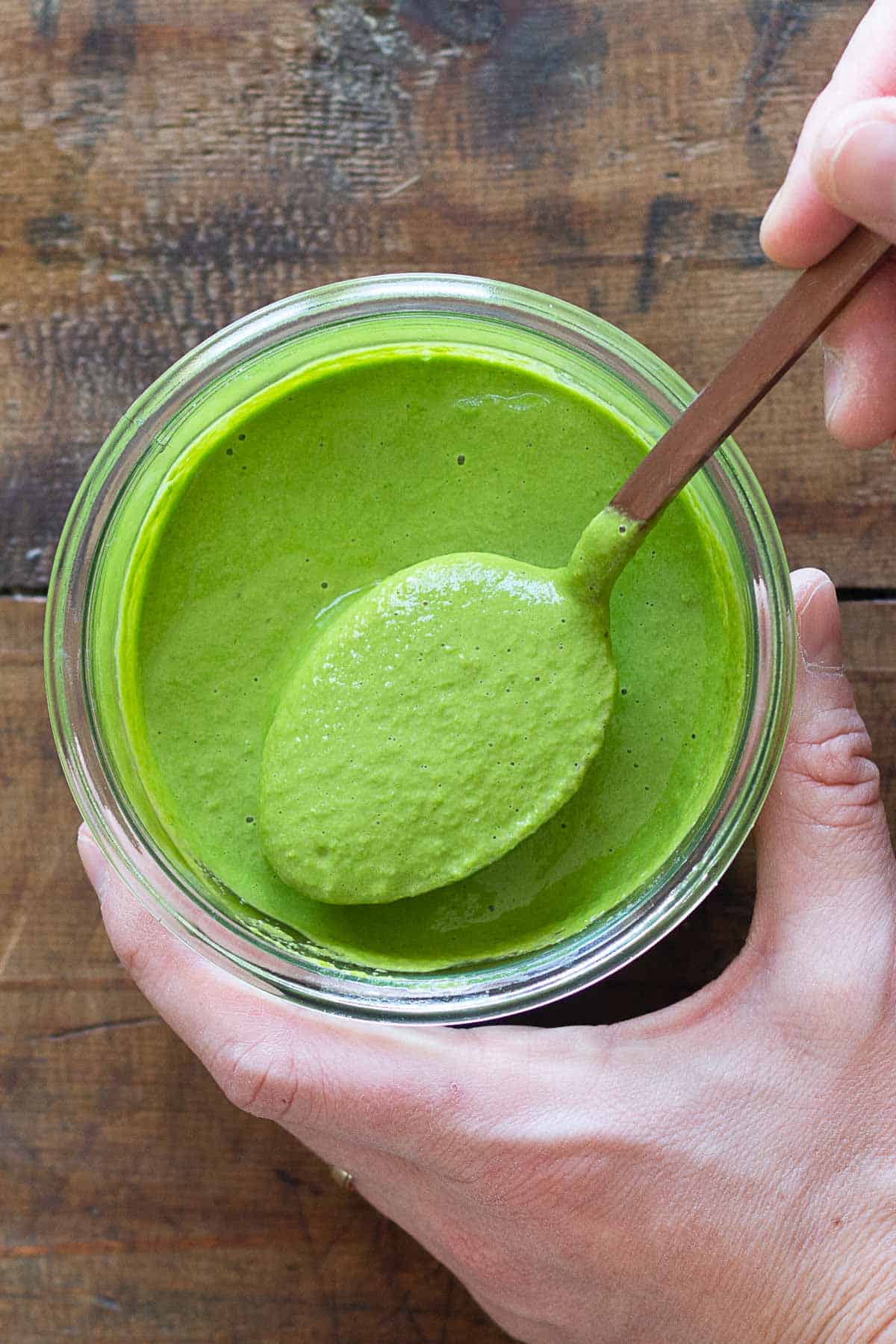 Green tahini sauce in jar and spoon lifting up green sauce.