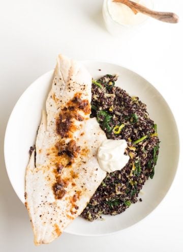 Top view of Haddock with Black Quinoa Risotto and creme fraiche on a white plate, and a cup with creme fraiche with a wooden spoon.