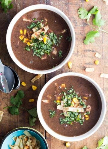 Two bowls of Mexican Black Bean Soup on a wooden board, two spoons and a small bowl of tortilla strips.