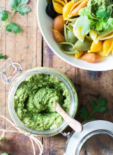 Top view of jar of Cilantro Pesto with a wooden spoon, and part of a bowl of rainbow shell pasta topped with Cilantro Pesto.