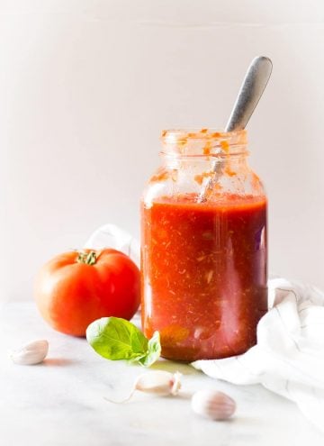 Roasted Garlic Tomato Sauce in a jar with a spoon, a fresh tomato, garlic cloves, and fresh basil leaves.