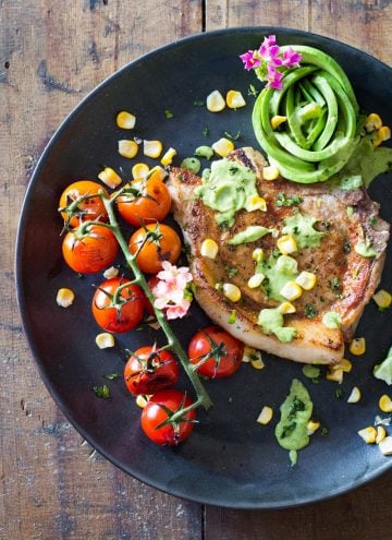 Mexican Pork Chops garnished with avocado cilantro sauce, corn kernels, cherry tomatoes and sliced lemon on a black plate.