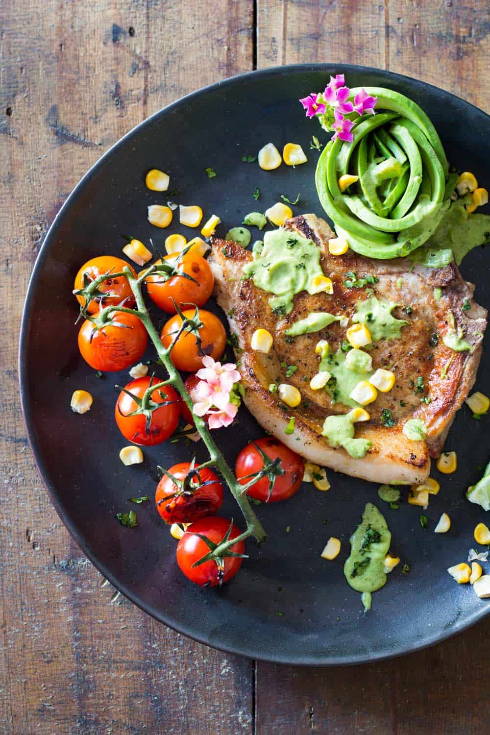 Mexican Pork Chops garnished with avocado cilantro sauce, corn kernels, cherry tomatoes and sliced lemon on a black plate.