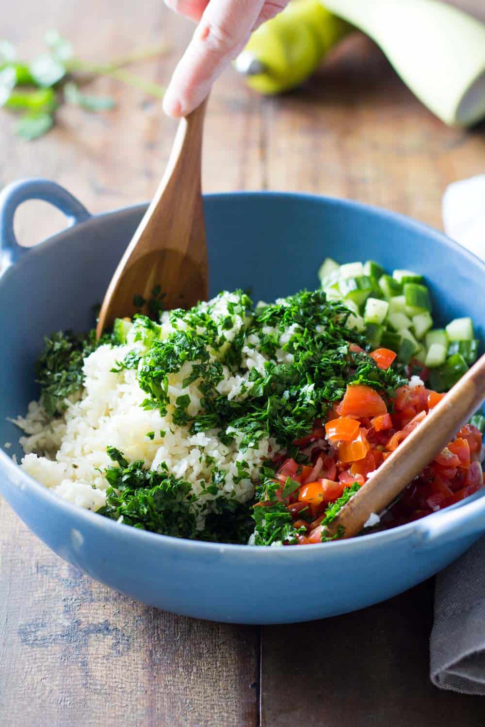 Cauliflower couscous tabbouleh salad being stirred with wooden spoons in a blue bowl. 