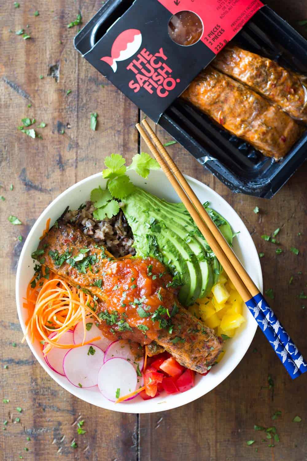Package of cooked salmon by The Saucy Fish Co. and a bowl of salmon poke salad with avocado, yellow and red pepper, radish, veggie noodles and wild rice, topped with sauce.