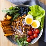 Bowl filled with lettuce, roasted sweet potato, roasted chickpeas, tomatoes, farro, red onion, blueberries, and a hard-boiled egg.