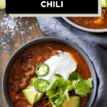 A bowl filled with chili con carne and topped with avocado, sour cream, jalapeno, and cilantro, with text overlay for Pinterest.