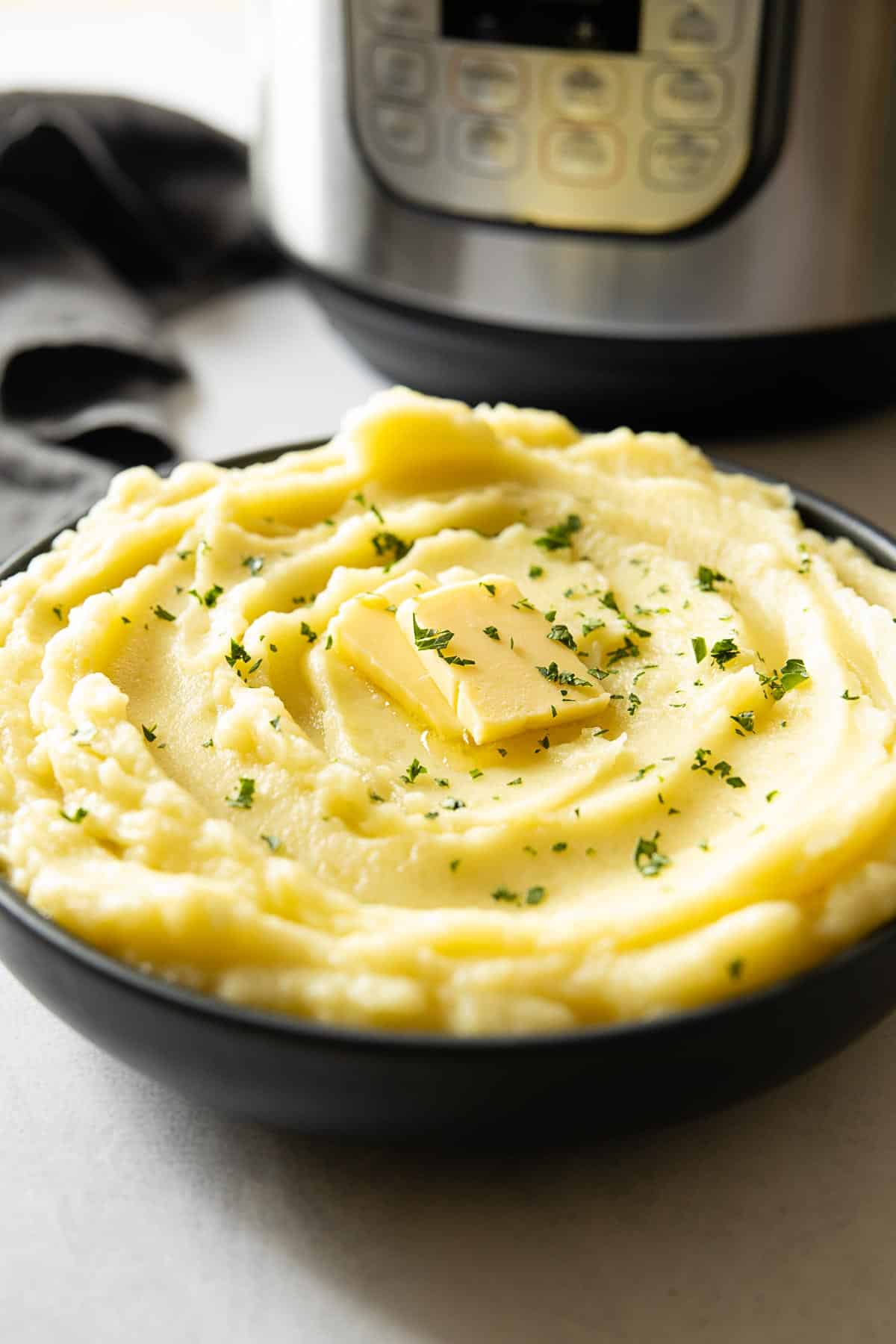 Creamy Mashed Potatoes in a bowl topped with butter and parsley, Instant Pot in background. Instant Pot Mashed Potatoes recipe