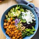 Lettuce, roasted sweet potato, avocado, onion, feta, cranberries in a blue salad bowl, dressing on the side.