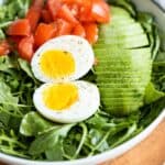 Arugula, hard-boiled egg, avocado, and tomato in a white salad bowl.