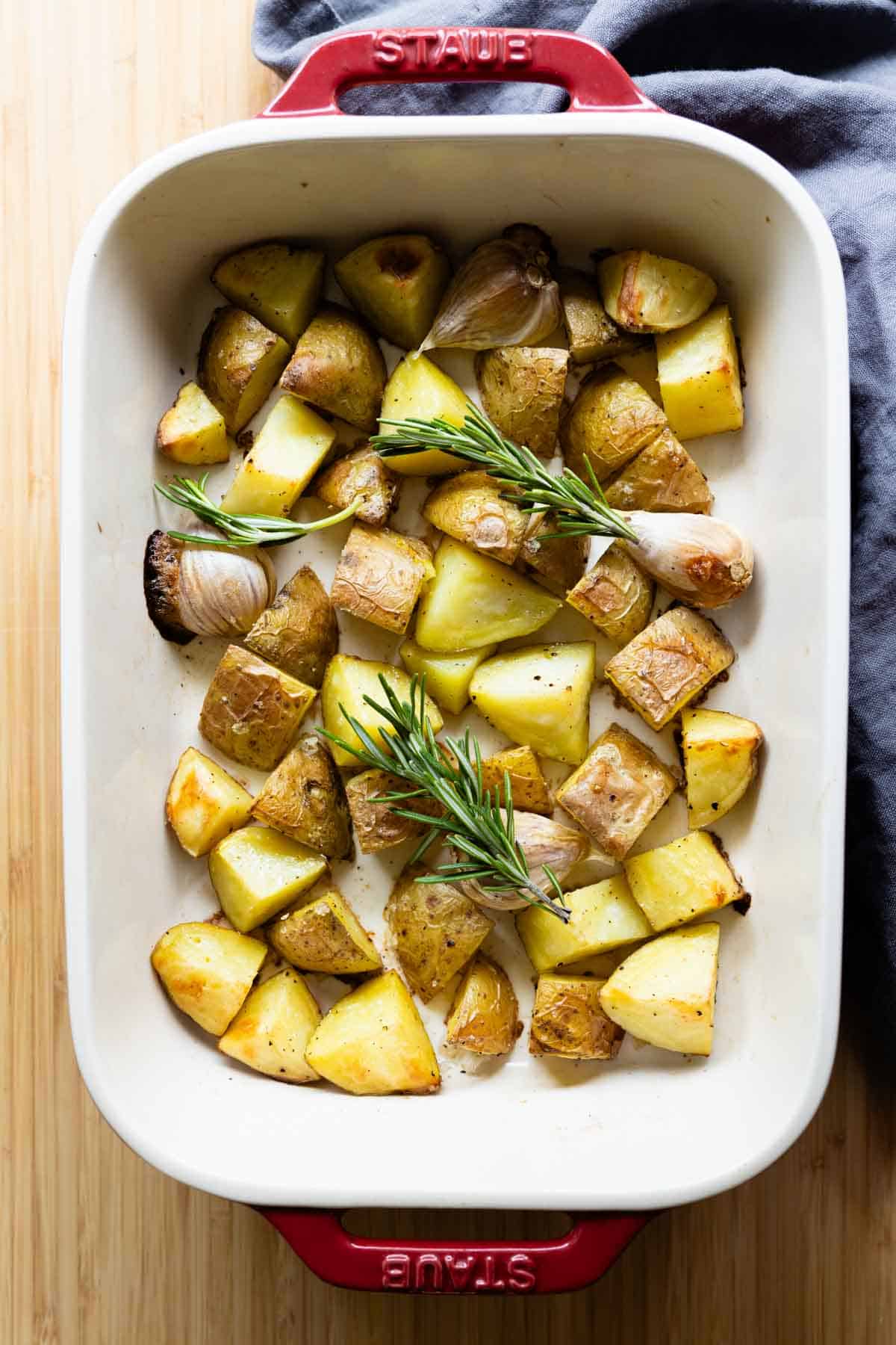 Whole garlic cloves, cut-up yellow potatoes, and rosemary in a white baking dish with red handles.